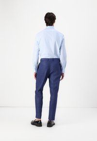 Ljusblå skjorta med långa ärmar, kombinerad med mörkblå skräddarsydda byxor och svarta skor, sedd bakifrån.