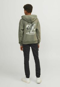 Sweat à capuche vert olive avec un design graphique blanc dans le dos, associé à un pantalon cargo noir et des baskets blanches.