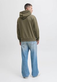 Man vanuit achteren bekeken, gekleed in een olijfgroene hoodie, lichtblauwe wijde jeans en witte sneakers, tegen een effen achtergrond.