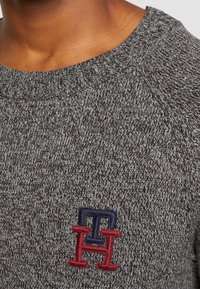 Grauer Strickpullover mit einem gerippten Kragen, der ein strukturiertes Muster aufweist und ein kleines gesticktes Logo in Marineblau und Rot auf der Brust trägt.
