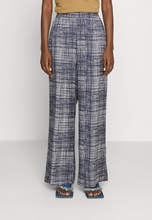 Persona que lleva pantalones de talle alto y pernera ancha con un patrón de rejilla azul marino y blanco y sandalias azules con borde deshilachado visibles en los pies.