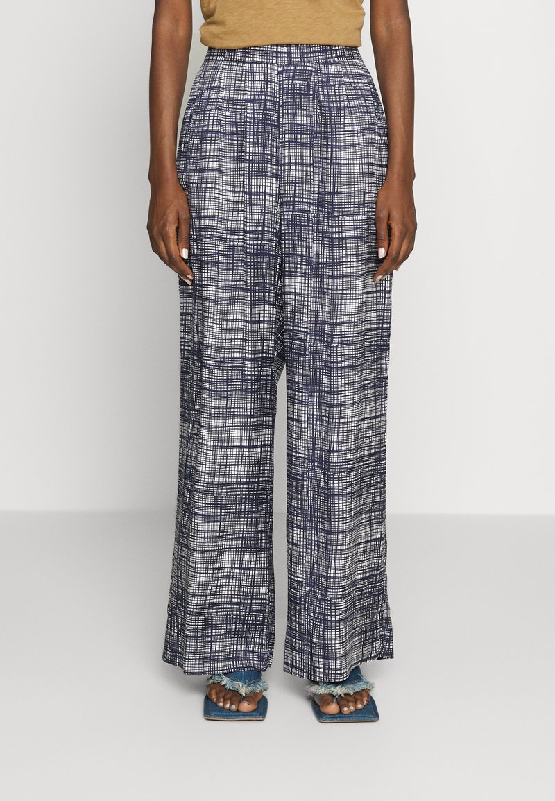 Personne portant un pantalon taille haute à jambes larges avec un motif à carreaux bleu marine et blanc et des sandales bleues à bords effilochés visibles aux pieds.