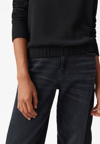Maglione nero con texture, dotato di orlo a coste, abbinato a jeans scuri. La vista frontale mostra una mano appoggiata sulla coscia, evidenziando i dettagli del tessuto.