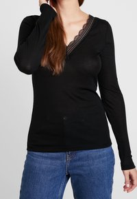 Černý dlouhý rukáv top s výstřihem ve tvaru V a krajkovým lemem. Tkanina vypadá jemně, je těsně přiléhavá v oblasti trupu, doplněná světle modrými džíny.