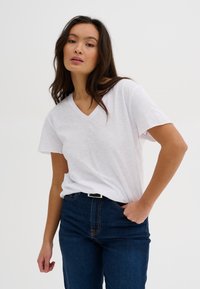 Biela V-krčný tričko s krátkymi rukávmi, vyrobené z mäkkej látky. Spárované s tmavomodrými džínsy s kontrastným oranžovým švom a čiernym opaskom.