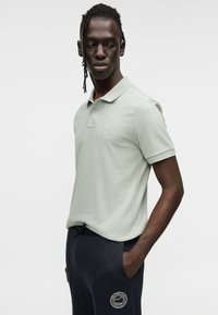 Ανοιχτό πράσινο πόλο μπλουζάκι με γιακά και κοντά μανίκια, με ένα μικρό λογότυπο. Συνδυάζεται με σκούρο μπλε παντελόνι με έμβλημα.