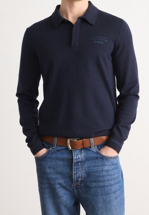 Mann trägt ein dunkelblaues Langarm-Poloshirt mit dem Logo „Porsche BOSS“, blaue Jeans und einen braunen Ledergürtel, Hände in den Taschen.