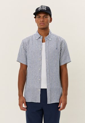 Chemise à manches courtes à boutons avec des rayures verticales bleues et blanches, en tissu léger, avec un logo sur la poitrine, portée par-dessus un t-shirt blanc.