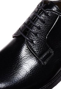 Chaussure en cuir noir avec une finition brillante, présentant un design classique à lacets, une tige texturée et une couture contrastante le long de la semelle.