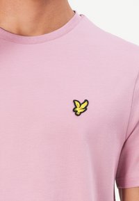 Mann iført en lys lilla T-skjorte med rund hals, med en liten gul fugl brodert på venstre bryst.