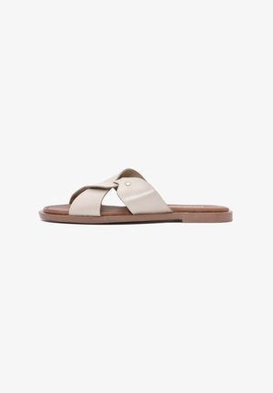 Beige slide sandal with crossed leather straps, flat brown sole, and a small metallic accent. Smooth texture, open design for breathability.