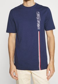Marinblå bomull t-shirt med en vit grafik till vänster och ett vertikalt rand i rött, vitt och grått ned längs sidan. Korta ärmar, rund hals.