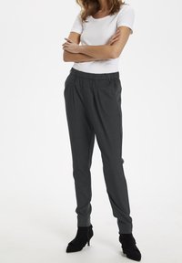 Grå skräddarsydda byxor gjorda av ett texturerat material, med ett elastiskt midjeband och sidofickor, tillsammans med svarta spetsiga klackar.