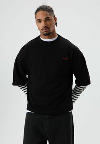 Chemise noire oversize à manches courtes avec un logo rouge, superposée sur un haut blanc à manches longues avec des rayures noires, associée à un pantalon noir.
