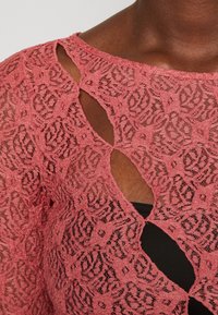 Top in pizzo rosa con aperture diagonali uniche sul petto, sopra una sottostruttura scura, che mostra un motivo in pizzo floreale testurizzato e la pelle.