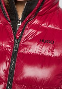 Rode pufferjack met glanzende afwerking, zwarte rits en "HUGO" logo. Heeft een gequilt ontwerp en hoge kraag voor warmte.