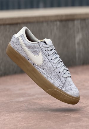 Grå tekstureret Nike-sneaker med hvid swoosh-logo og sål af gummigummi, vist svævende over en betonoverflade.