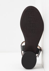 Mustat sandaalit, joissa on litteä pohja, sileä nahkapinta, nilkkahihna ja kohokuvioitu teksti, joka lukee "Valmistettu rakkaudella Espanjassa."