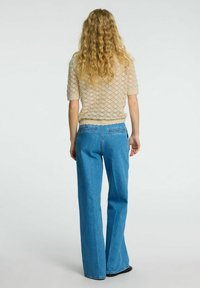Beige pletený top s texturovaným diamantovým vzorem, v kombinaci s vysokými modrými džíny s rozšířenými nohavicemi, které ukazují uvolněnou siluetu.