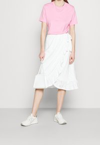 Rosa Baumwoll-T-Shirt, weiße Lochstickerei Wickelröcke mit Rüschensaum und weiße Handtasche; getragen mit hellen Sportschuhen.