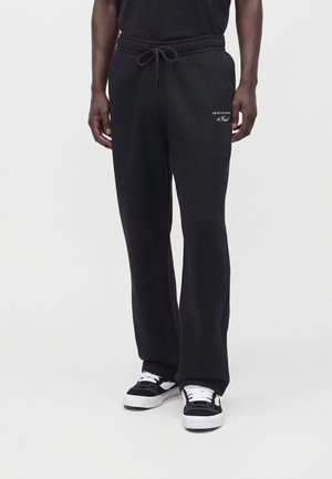 Uomo con pantaloni della tuta neri con cordoncino e sneakers nere e bianche, in piedi davanti a uno sfondo chiaro e uniforme.