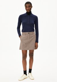 Femme portant un polo à manches longues bleu marine, une jupe en laine marron, des chaussettes noires et des chaussures blanches à lacets, se tenant devant un fond blanc.