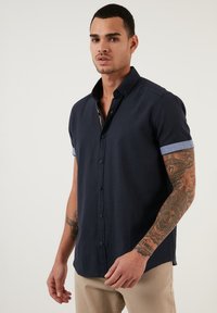 Cămașă de bumbac bleumarin cu mânecă scurtă, cu guler tip button-down și manșete de un albastru deschis; realizată dintr-un material moale; are butoane în față și detalii subtile de cusătură.