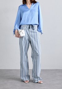 Světle modrá blůza s límečkem a dlouhými rukávy, spárována s volnými pruhovanými kalhotami ve světle modré a bílé, držící malou bílou tašku.