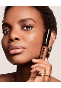 Prodotto labbra marrone in un tubo trasparente con tappo nero, tenuto accanto a un primo piano di un volto con pelle liscia e trucco naturale.