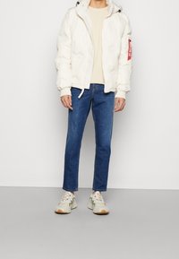 Weißer Pufferjacke mit Kapuze und gerippten Bündchen, beigem Rundhals-Shirt, blauen Slim-Fit-Jeans und grauen Sneakers mit grünen Akzenten.