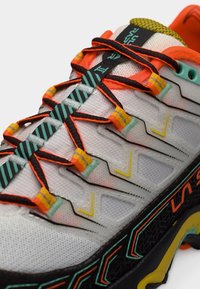 Λευκό αθλητικό παπούτσι με δίχτυ, με μαύρες, πορτοκαλί και κίτρινες λεπτομέρειες. Διαθέτει πολύχρωμα κορδόνια και υφή στη σόλα για καλύτερη πρόσφυση.