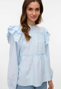 Blusa azzurro chiaro in cotone con dettagli arricciati sulle spalle, vita arricciata e maniche lunghe. Texture liscia con un leggero lucido.