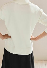 Weißer Strickpullover mit hohem Kragen, geripptem Saum und übergroßen Ärmeln. Die Rückseite hat eine glatte Textur und eine lockere Passform.