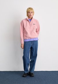 Een persoon staat met een roze fleece pull-over, een lichtpaarse shirt, losse blauwe jeans en zwarte schoenen tegen een plain witte muur.