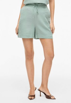 Kvinne iført lysegrønne høytlivsshorts med snor og brune åpen-tå hælsko, står med hendene i lommene.