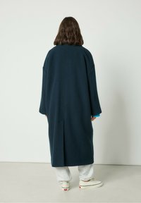 Dlhý námornícky vlnený kabát s voľným strihom, spustenými ramenami a jedným zadným rozparkom. V kombinácii so šedými teplákmi a plátennými topánkami.