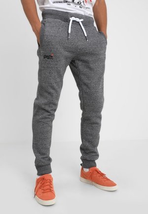 Personne portant un pantalon de survêtement gris avec cordon de serrage et poches latérales, ainsi que des baskets orange, debout devant un fond clair uni.