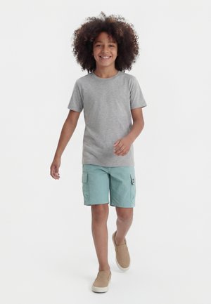 Χαμογελαστό παιδί με σγουρά μαλλιά που περπατά μπροστά, φορώντας γκρι κοντομάνικη μπλούζα, τιρκουάζ παντελόνι cargo και μπεζ παπούτσια slip-on.