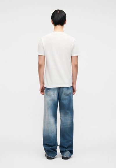 Λευκό μπλουζάκι με κοντά μανίκια σε συνδυασμό με φαρδιά, ανοιχτόχρωμα τζιν παντελόνια, με ξεθωριασμένο πλύσιμο και διακριτικές φθορές.