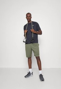 Asmos sportiniais drabužiais, asmuo vilki tamsiai mėlyną trumpą rankovę marškinėlį ir alyvuogių žalumos šortus, laiko pasipriešinimo juostas. Pilki batai ir balti kojinės užbaigia išvaizdą.