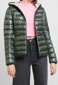 Mørkegrøn pufferjakke med en blank overflade og kviltet design, med lynlås foran og hætte med grå for. Båret over en lyserød top.