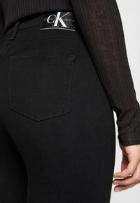 Närbild av svarta Calvin Klein-jeans med baksida och logopatch, ihop med en svart ribbad långärmad topp och handen vilande vid sidan.