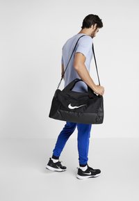 Czarna torba sportowa Nike z wyraźnym białym logo, wyposażona w podwójne uchwyty, pasek na ramię oraz teksturowany materiał. Model ma na sobie niebieskie spodnie i czarne buty.