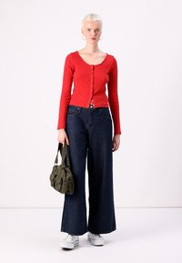 Cardigan rouge à rayures et à manches longues avec fermeture à boutons, associé à un jean en denim noir à jambes larges. Porte un sac en tissu vert. Baskets blanches.