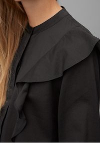 Black blouse featuring a ruffled collar and placket, crafted from smooth fabric with a matte texture. Displayed on a neutral background.