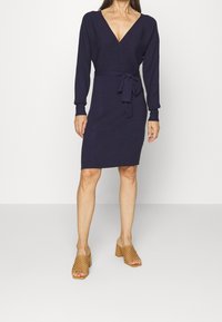 Abito avvolgente navy in tessuto a costine con maniche lunghe, vita cinturata e scollatura a V. Abbinato a mule con tacco aperto color sabbia.