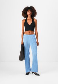 Crop top noir à côtes avec un décolleté en V profond, associé à un pantalon large plissé bleu clair. Le mannequin porte un tote bag marron foncé. Talons noirs.
