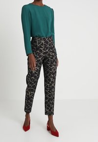 Žena stojí v azurově modré blůze s dlouhými rukávy a vzorovaných černých kalhotách, obutá do červených podpatků, na jednobarevném pozadí.