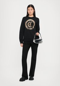 Schwarzer Sweatshirt mit einem kreisförmigen, goldmusterigen Logo, kombiniert mit schwarzen Schlaghosen und einer kleinen, metallisch silbernen Handtasche.