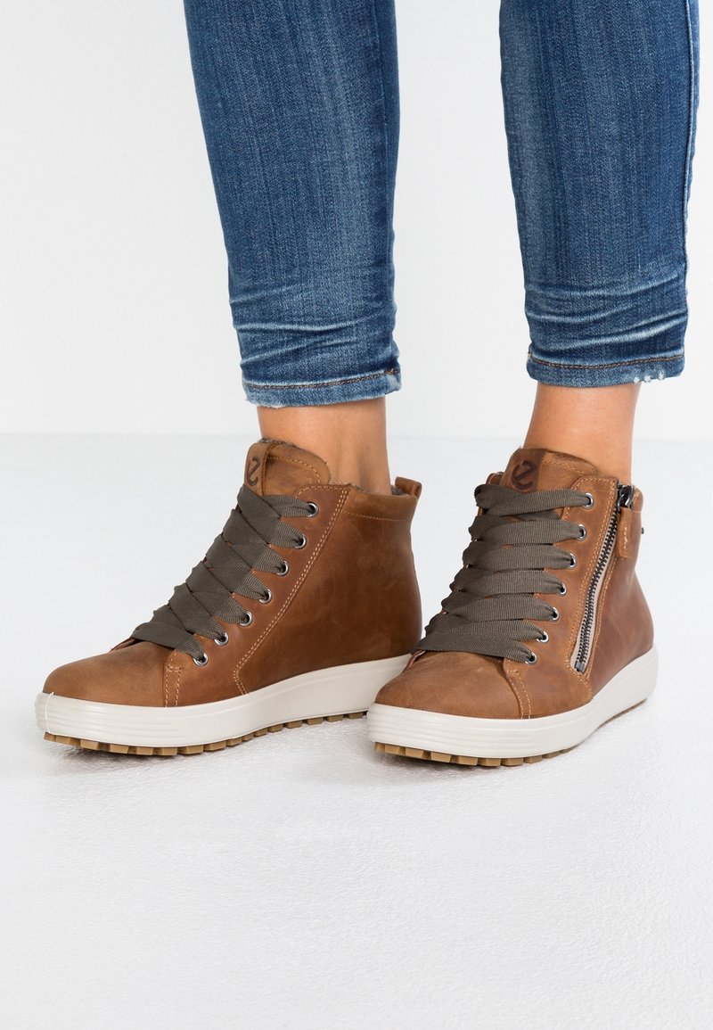 Baskets montantes en cuir marron avec de gros lacets gris et des fermetures éclair latérales, portées avec un jean bleu retroussé sur un sol blanc.